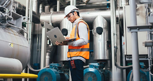 An engineer on a plant floor consults their laptop.